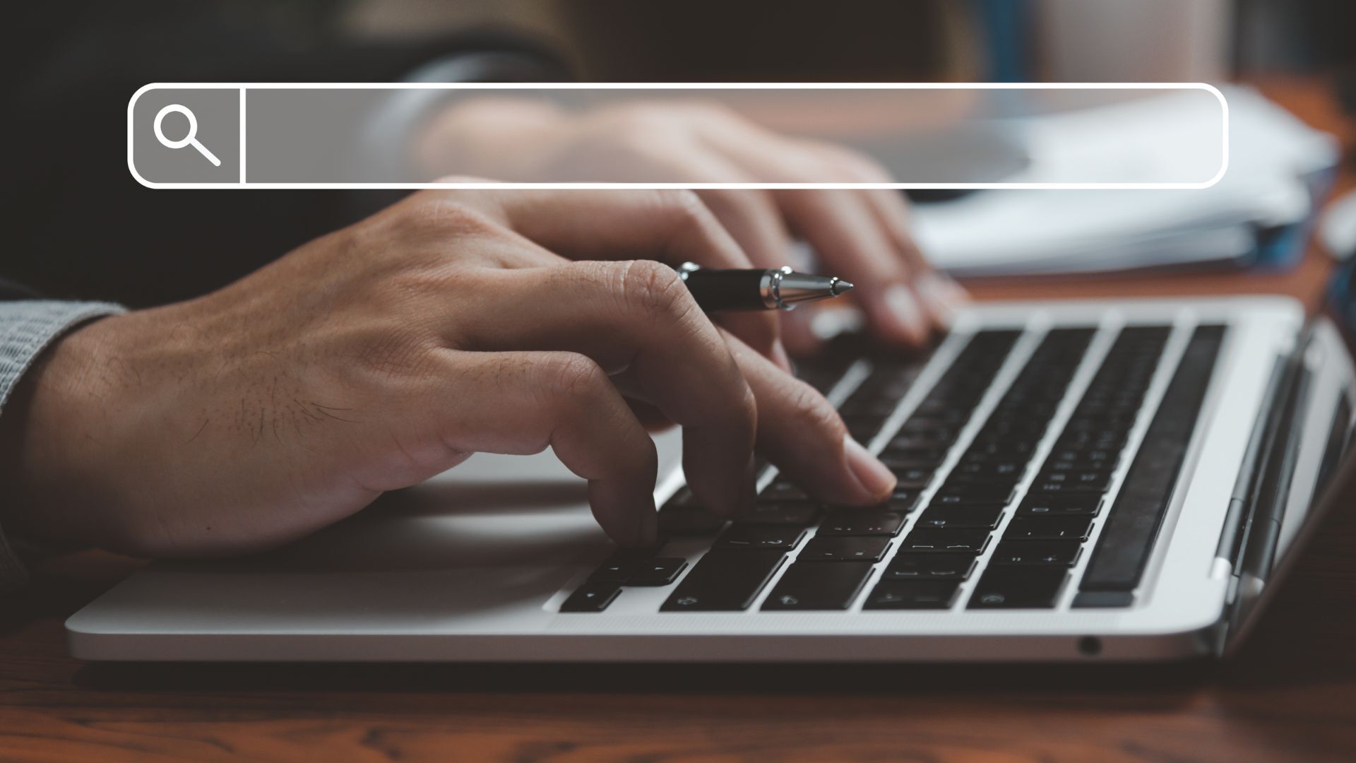A close up image of a person typing on a silver computer with black buttons. An empty search engine query is overlaid on the photo.